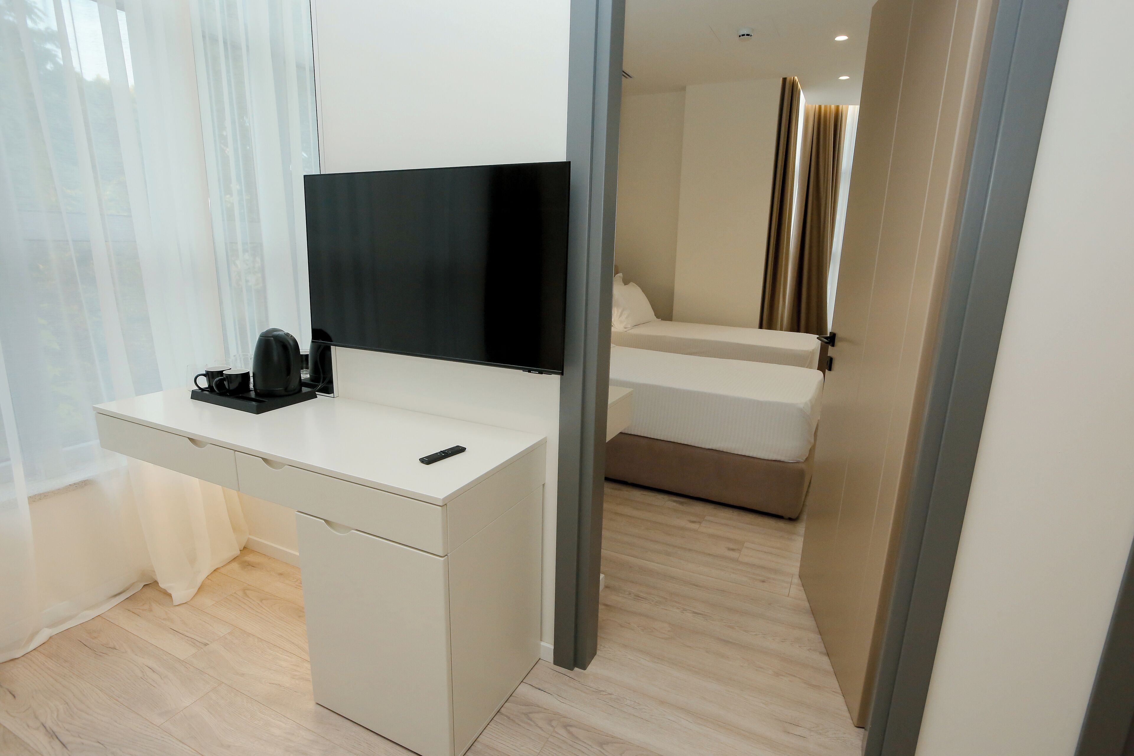 Quarto de hotel moderno com uma escrivaninha branca, televisão de tela plana, cafeteira e uma cama com cabeceira bege.