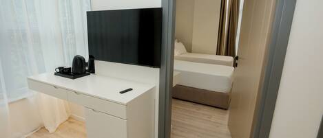A modern hotel room with a white desk, a flat-screen TV, a coffee maker, and a bed with a beige headboard.
