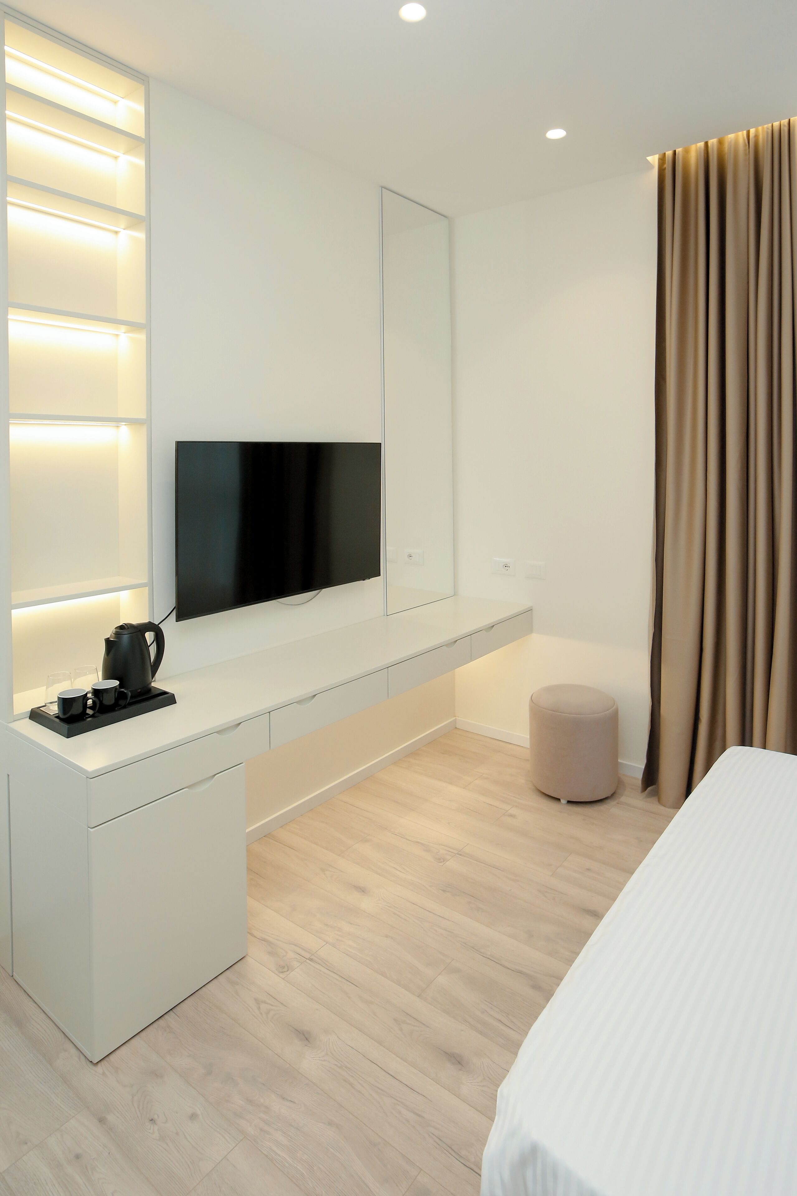 A modern hotel room with a white console table, a flat-screen TV, a bedside table, a round ottoman, and a large window with curtains.