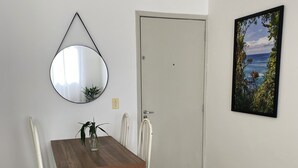 A dining area with a wooden table and white chairs, a round mirror on the wall, a door, and a framed picture of a landscape.