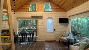 Interior de una cabaña con techo de madera, dos ventanas, un comedor y una sala de estar con sofá y sillones.