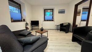 Een moderne woonkamer met een bank, een salontafel en een televisie. Twee ramen met zonwering, een klein tafeltje met een plant en een schilderij aan de muur.