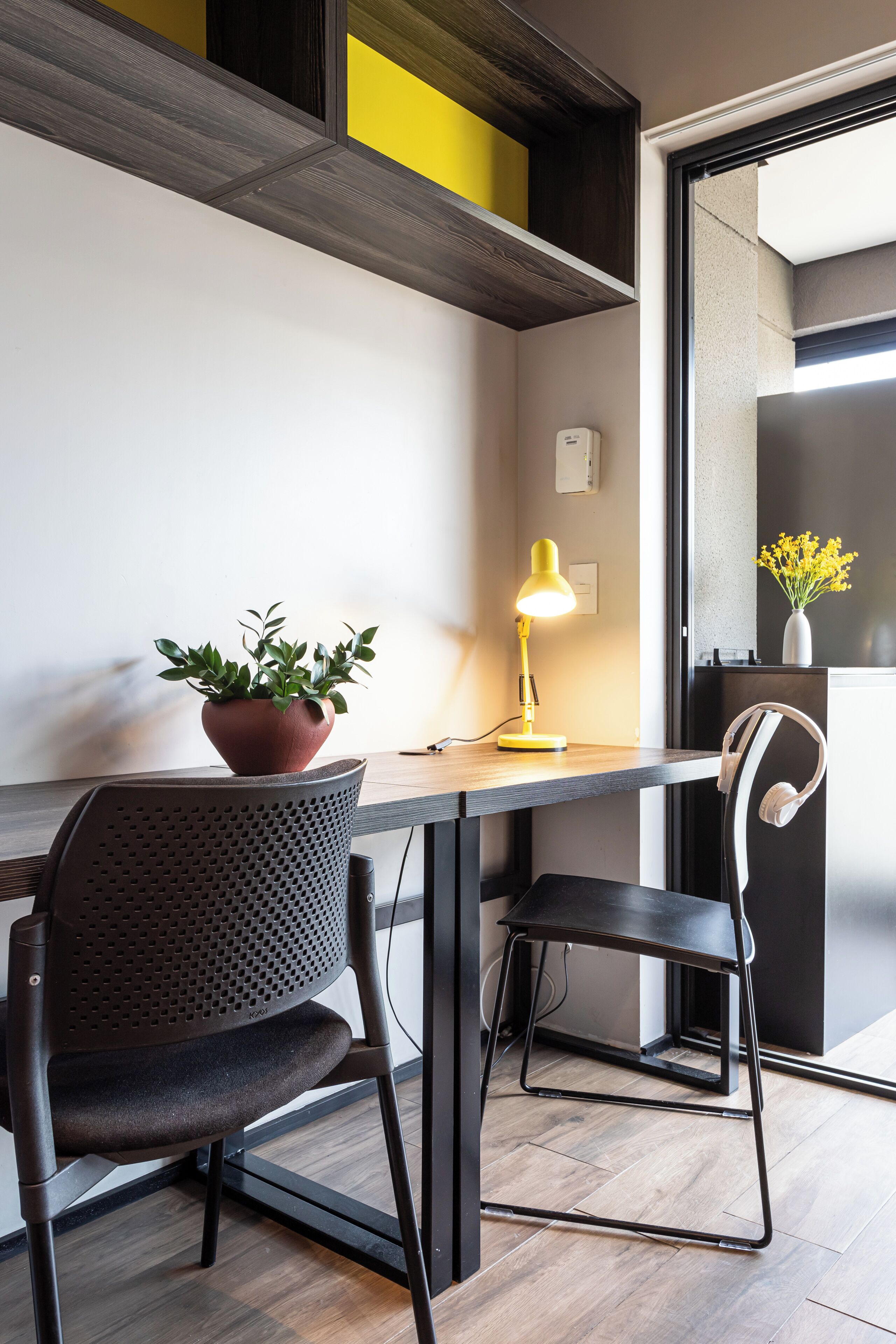 A modern workspace with a desk, two chairs, a potted plant, a yellow desk lamp, and a vase with flowers.