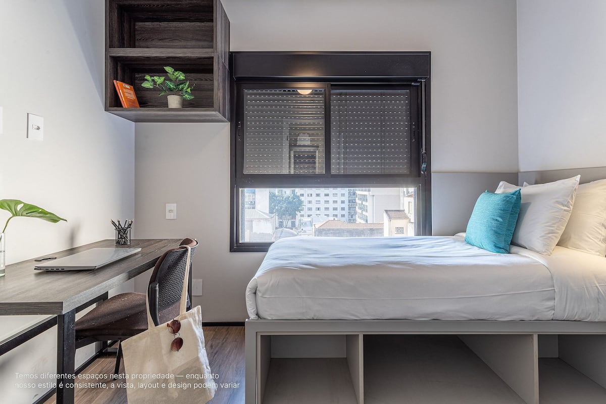 A bedroom with a bed, a desk, and a window with blinds.