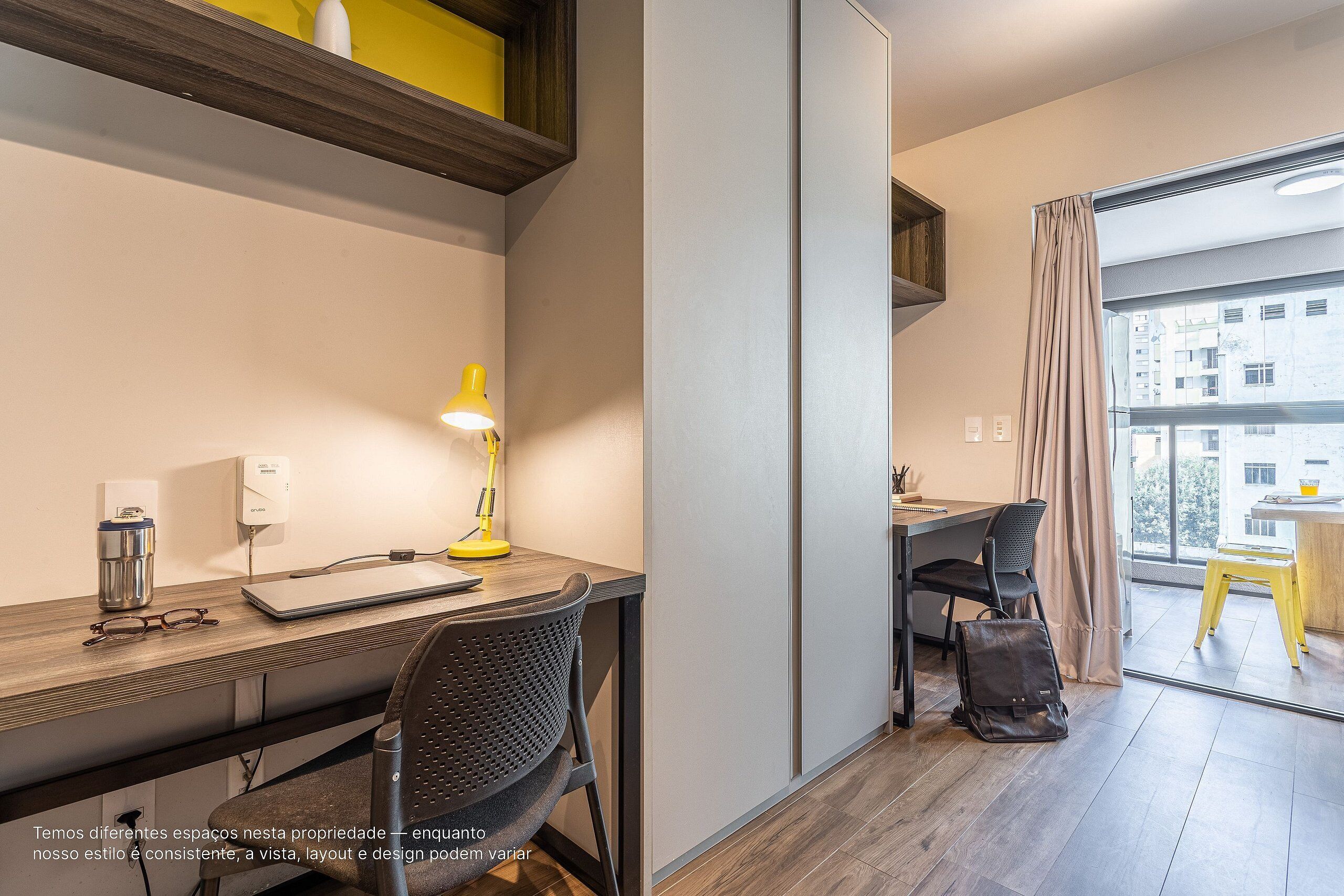 A modern hotel room with a desk, chair, lamp, and a view of the city through large windows.