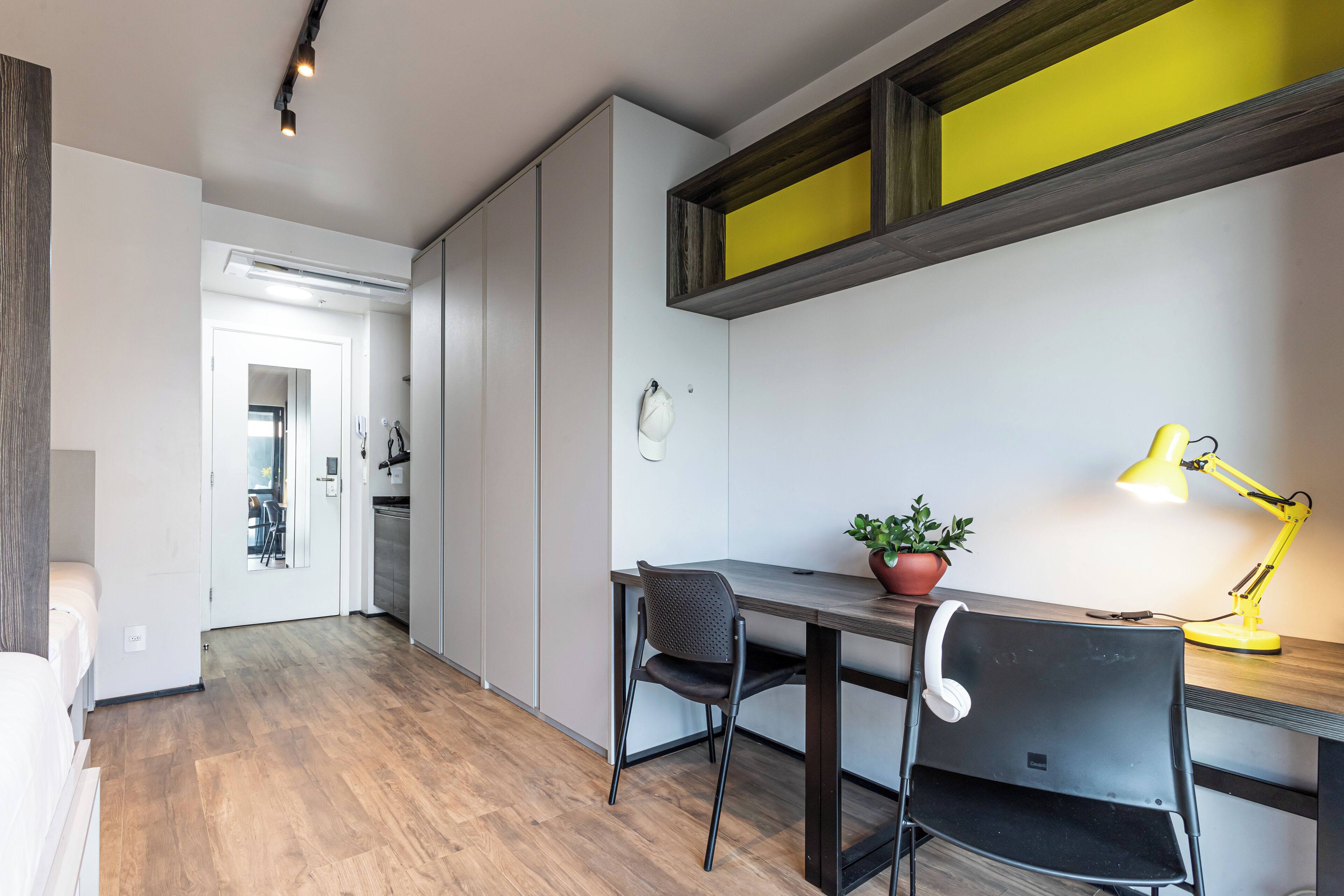 A modern room with a wooden floor, a desk with a chair, a potted plant, and a yellow desk lamp.