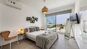 A modern bedroom with a large bed, a wicker chair, a bedside table with a lamp, a wall-mounted air conditioner, and a large window with a view. - Protaras Bay View Villa 11 (Paralimni)