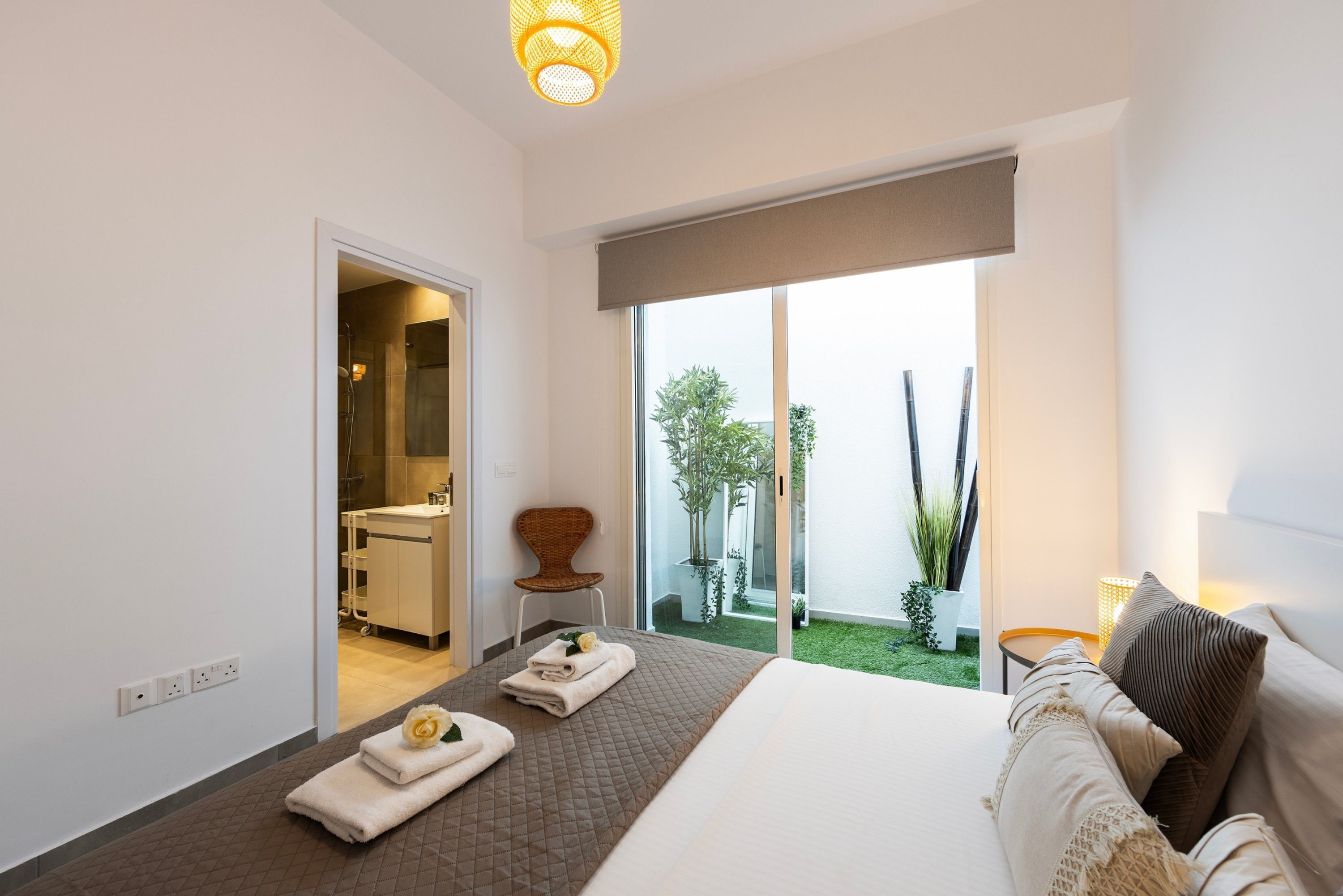 Un dormitorio moderno con una cama grande, un baño visible a través de una puerta abierta y vistas a un espacio verde exterior.