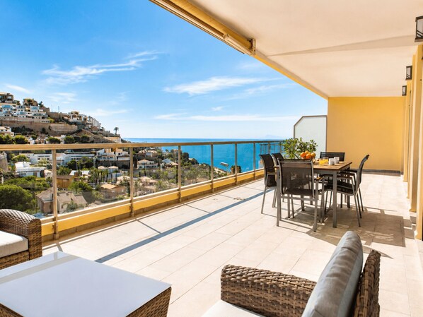A balcony with a table and chairs, overlooking a coastal town and the sea.