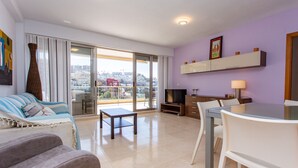 A modern living room with a sofa, a coffee table, a dining table with chairs, a TV, and a balcony with a city view.