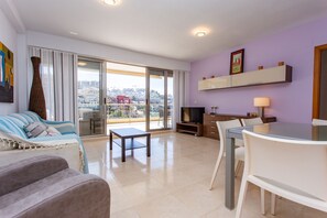A modern living room with a sofa, a coffee table, a dining table with chairs, a TV, and a balcony with a city view.