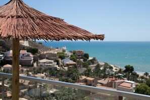 A balcony with a straw umbrella overlooking a coastal town with buildings and the sea.