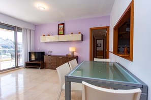 A modern living room with a glass-top dining table, a flat-screen TV, and a sliding glass door leading to a balcony.