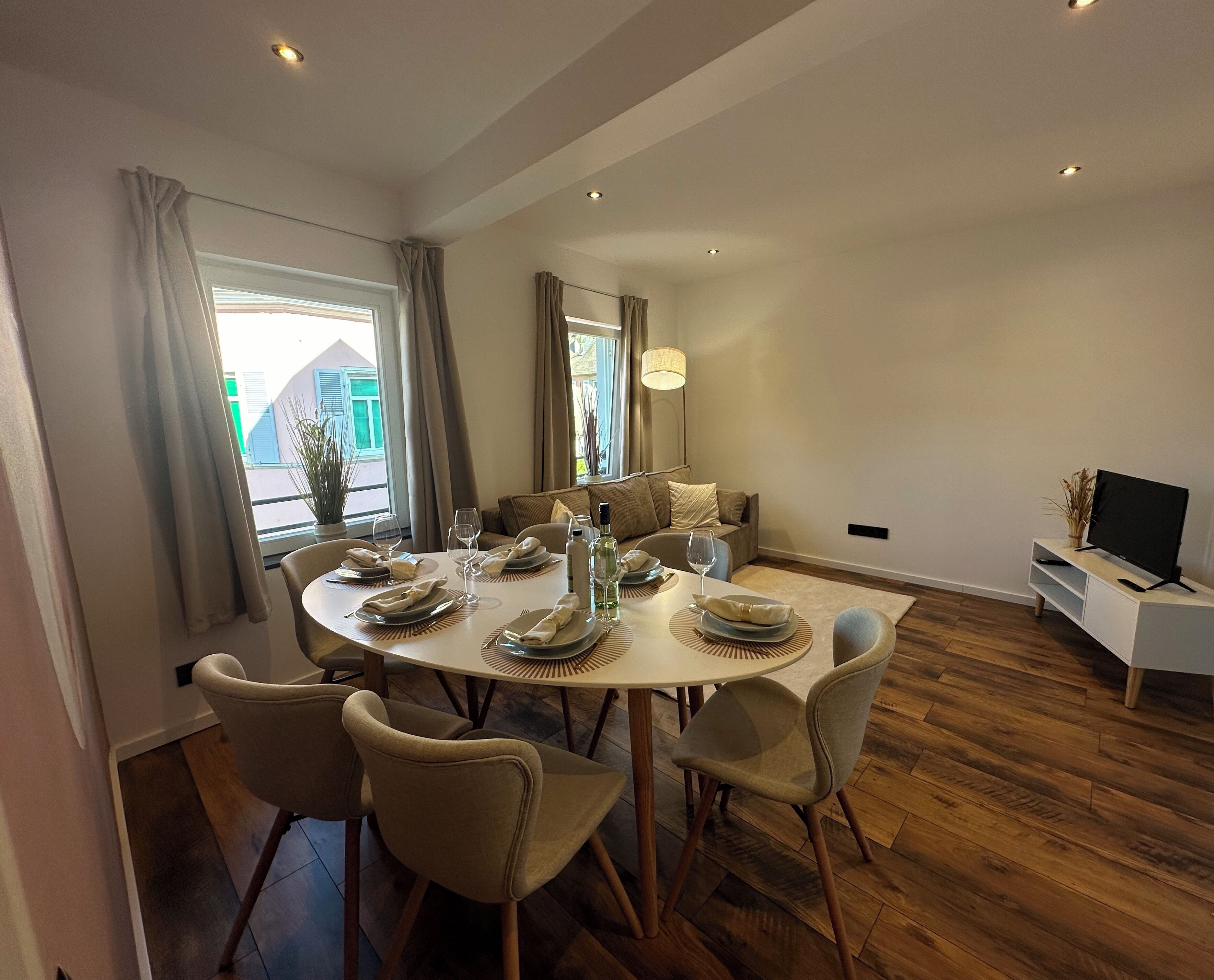 Ein Essbereich mit einem runden Tisch, gedeckt für vier Personen, umgeben von vier Stühlen. Im Raum befindet sich außerdem eine Couch und ein TV-Schrank.