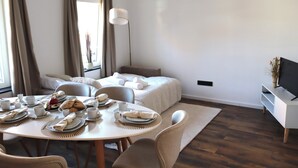 Una habitación de hotel moderna con zona de comedor, cama, televisor y un gran ventanal con cortinas.