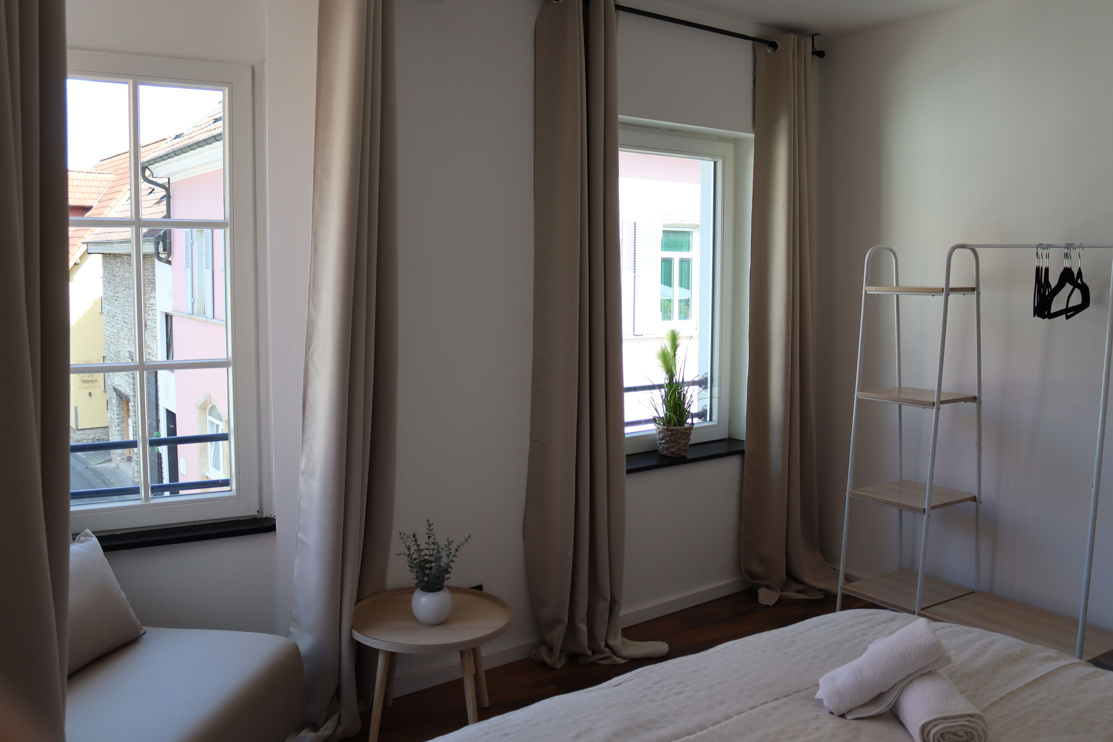 Ein Schlafzimmer mit einem Bett, einem Fenster mit Vorhängen, einem kleinen Tisch mit Pflanze und einem Metallregal mit Kleidung.