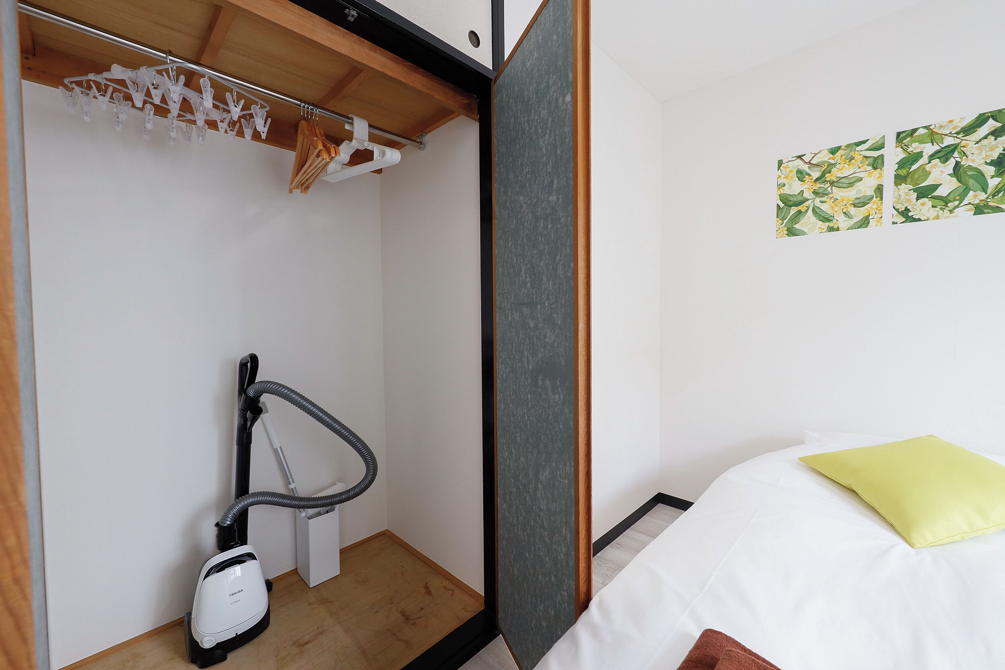 A compact room with a bed, a vacuum cleaner, a wooden shelf with hanging clothes, and a wall-mounted artwork.