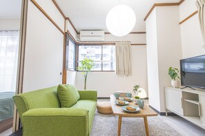 A modern living room with a green sofa, a wooden coffee table, a TV, and a window with a view of buildings. - Solaria Tokiwa (Tokyo)