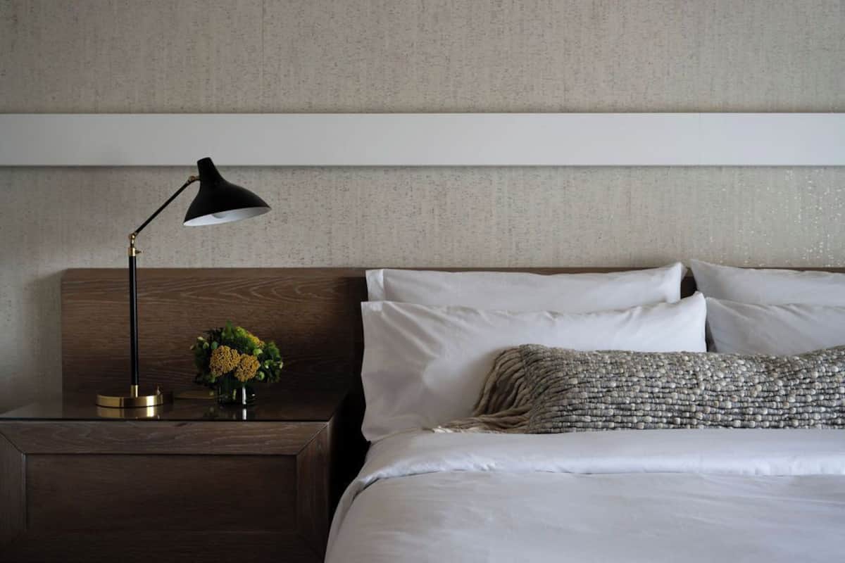 Un letto rifatto con lenzuola bianche e un cuscino decorativo, un comodino con una lampada nera e un piccolo vaso con fiori gialli.