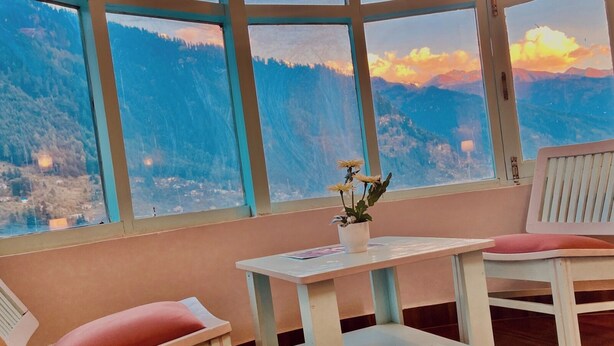 A room with a large window offering a mountain view, a small table with a potted plant, and two white chairs with red cushions. - Hotel Hadimba palace  (Manali)