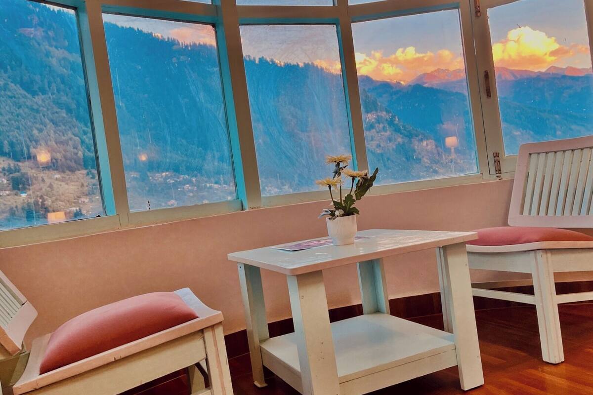 A room with a large window offering a mountain view, a small table with a potted plant, and two white chairs with red cushions.