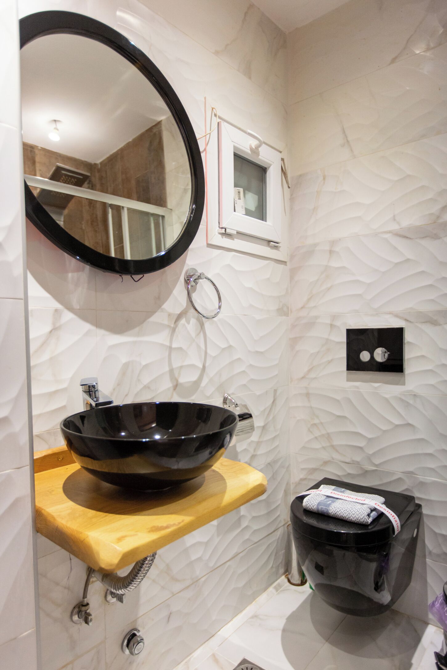 A bathroom with a round mirror, a black vessel sink, a wooden shelf, a toilet, and a textured wall.