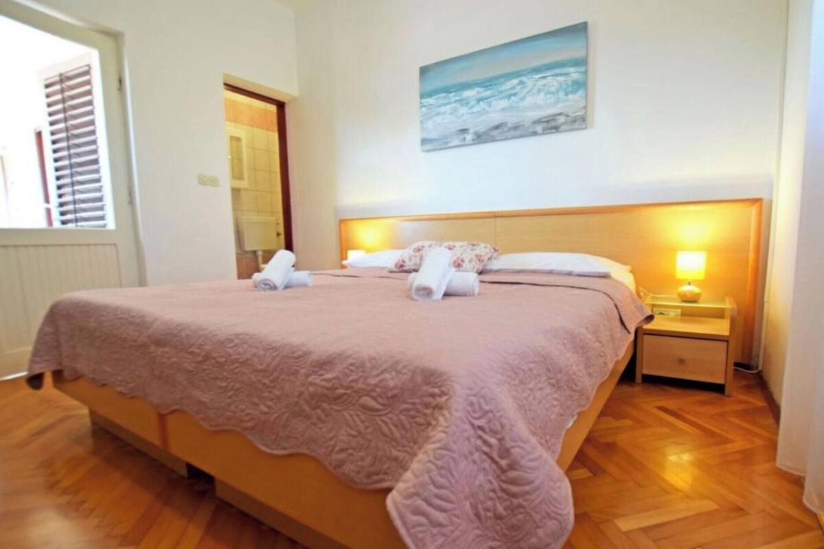 A neatly made bed with a pink blanket, a painting of a beach scene on the wall, and wooden flooring.