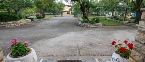A paved driveway leading to a building with a stone wall on one side and a metal fence on the other. There are flower pots with pink and red flowers on either side of the driveway.