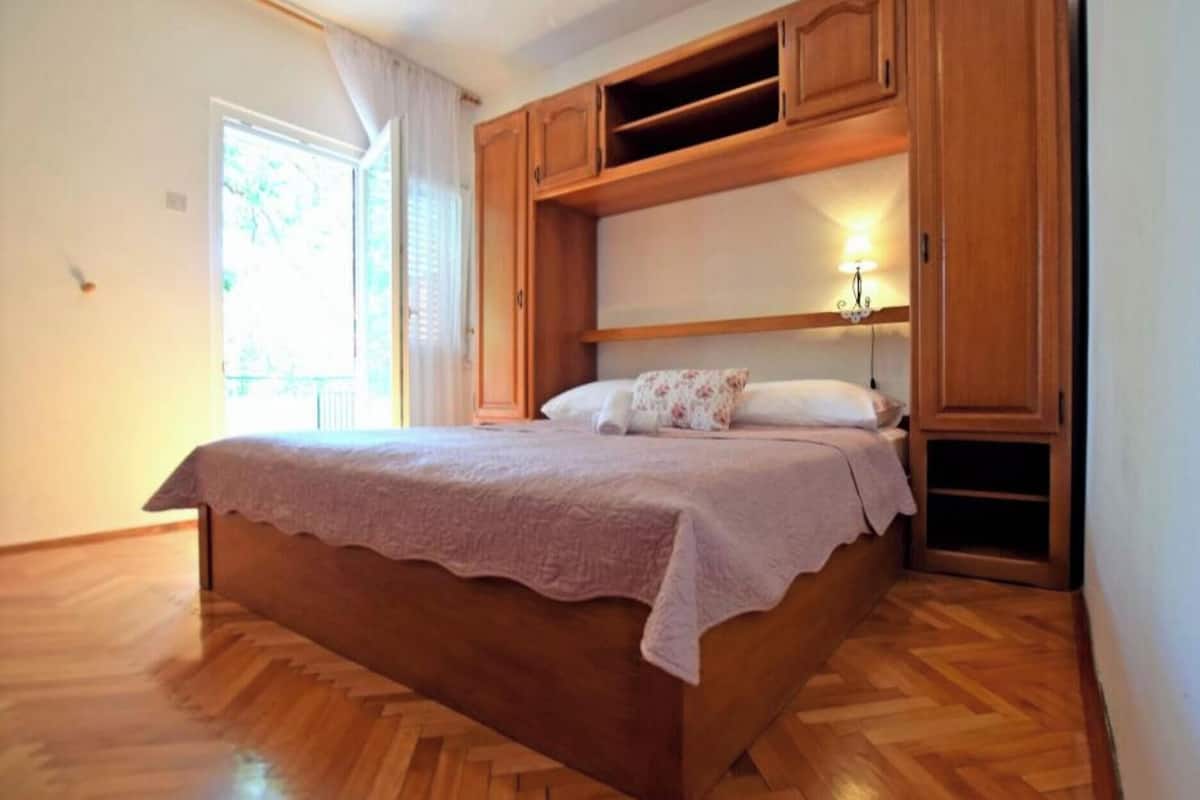 A bedroom with a wooden bed, a window with curtains, and a built-in shelf above the bed.
