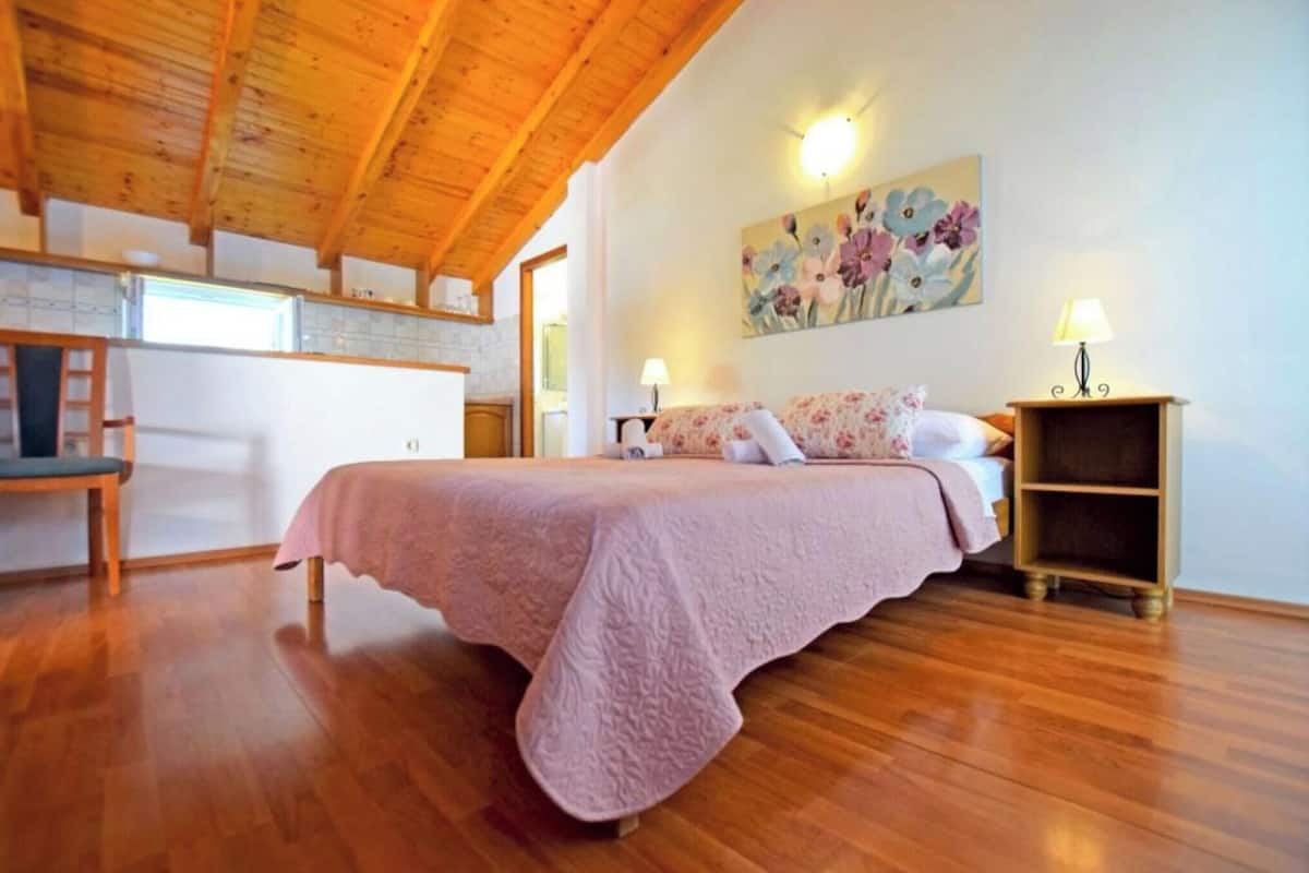 A bedroom with a wooden ceiling, a bed with a pink bedspread, a nightstand with a lamp, and a painting on the wall.
