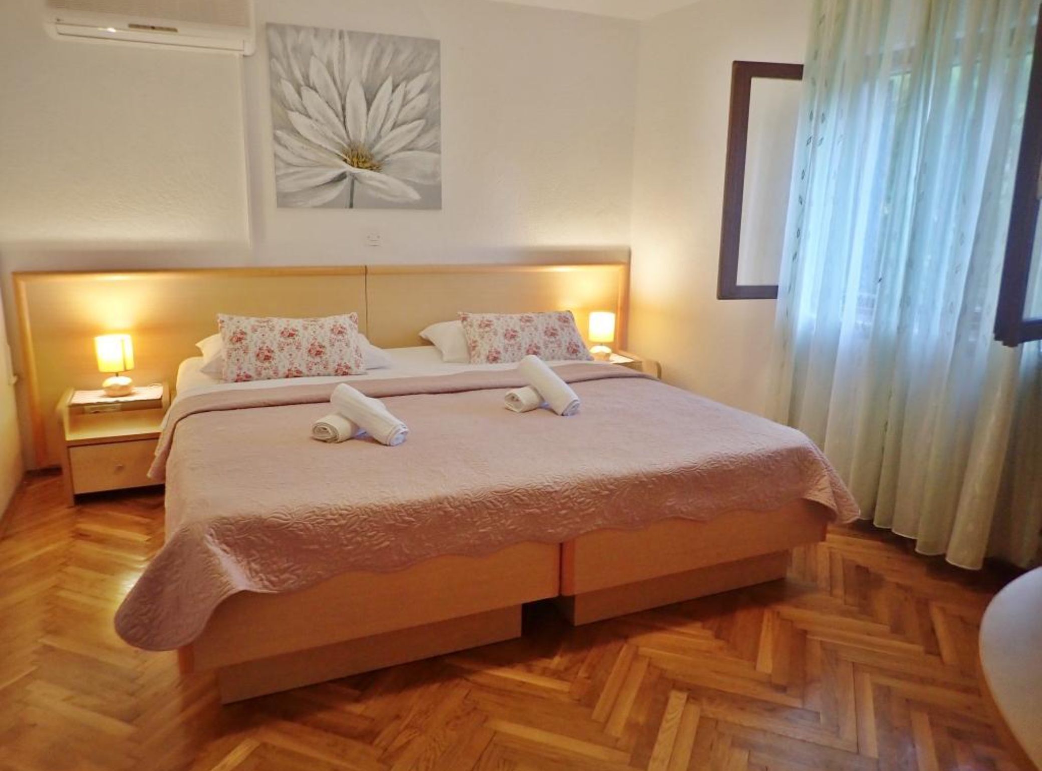 A hotel room with a large bed, two bedside tables with lamps, a floral painting on the wall, and a window with curtains.