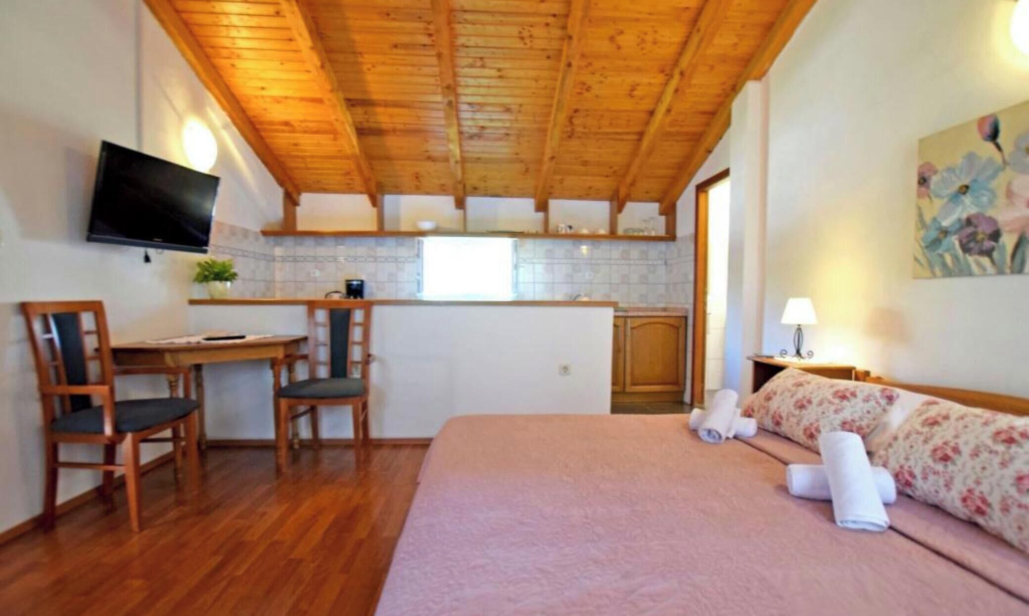 A bedroom with a wooden ceiling, a bed with pink bedding, a desk with two chairs, a TV mounted on the wall, and a kitchen area in the background.