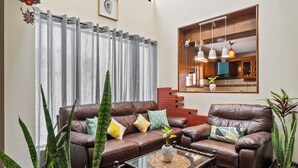A living room with a brown leather sofa, a glass coffee table, and a large window with curtains. - 🌳 Namma Woodland Villa | Golf Course Views (Bengaluru)