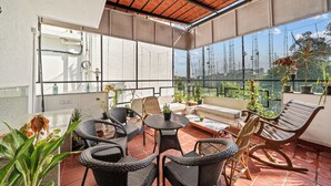 A covered outdoor patio with wicker furniture, potted plants, and a view of the cityscape. - 🌳 Namma Woodland Villa | Golf Course Views (Bengaluru)