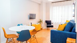 Un salon moderne avec un canapé bleu, des fauteuils jaunes et bleus, une table blanche et un téléviseur fixé au mur.
