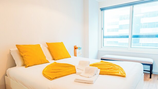 Un lit bien fait, avec du linge de lit blanc et deux coussins jaune vif, une petite table de chevet avec une bouteille, et un banc près de la fenêtre.