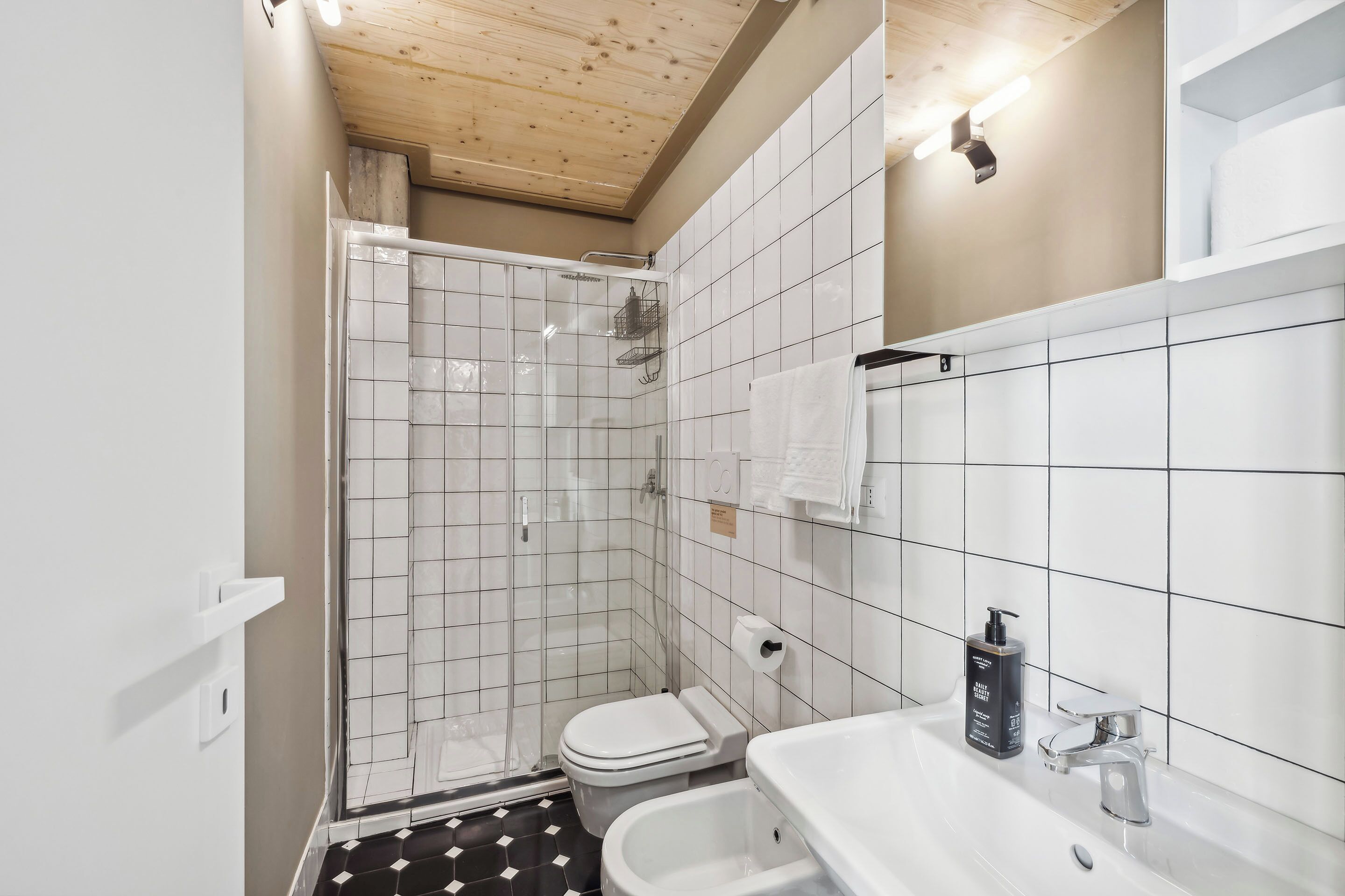 Un bagno moderno con doccia, water e vasca, rivestito di piastrelle bianche e con soffitto in legno.