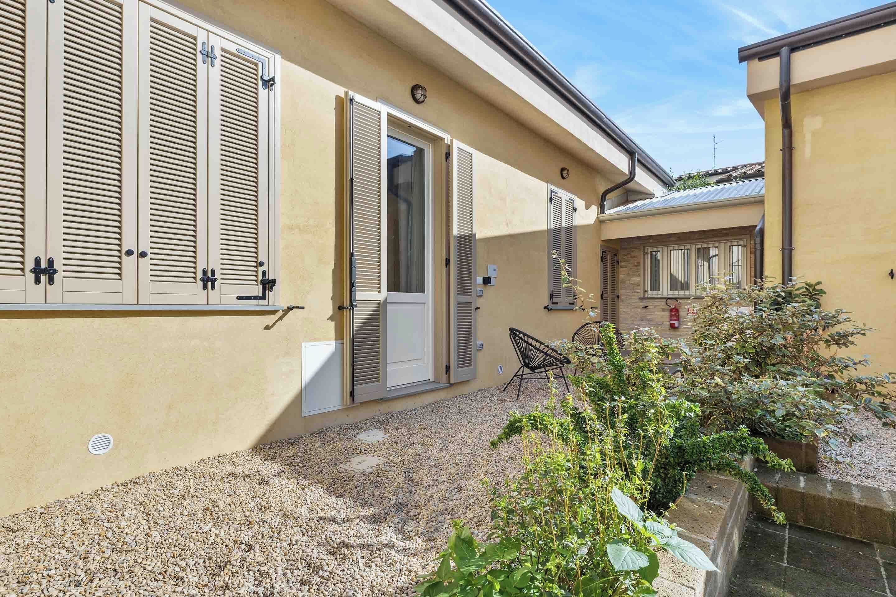 Un edificio con facciata color beige, vialetto in ghiaia e porta che si apre su un patio coperto.