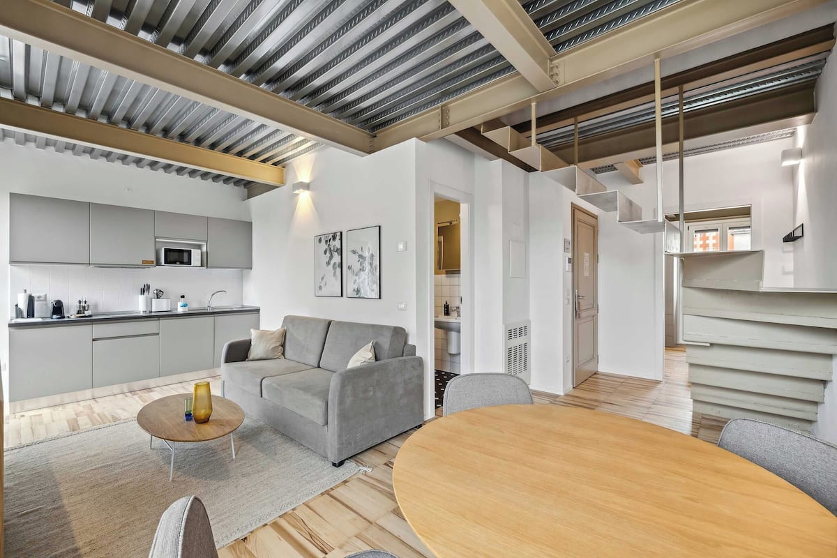 A modern kitchen with a built-in microwave and stainless steel appliances, a grey sofa with a coffee table, and a wooden dining table in a bright living area with a high ceiling and exposed beams.