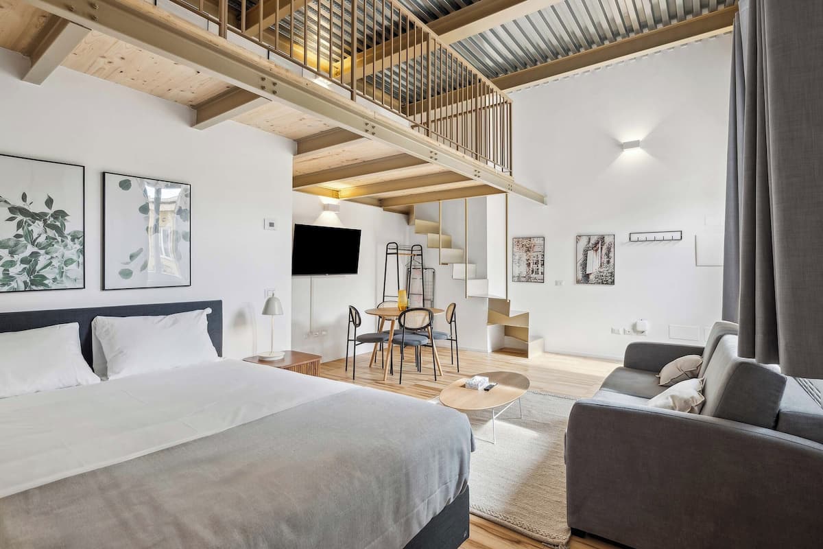 A modern hotel room with a large bed, a sofa, a dining area, and a staircase leading to an upper level.