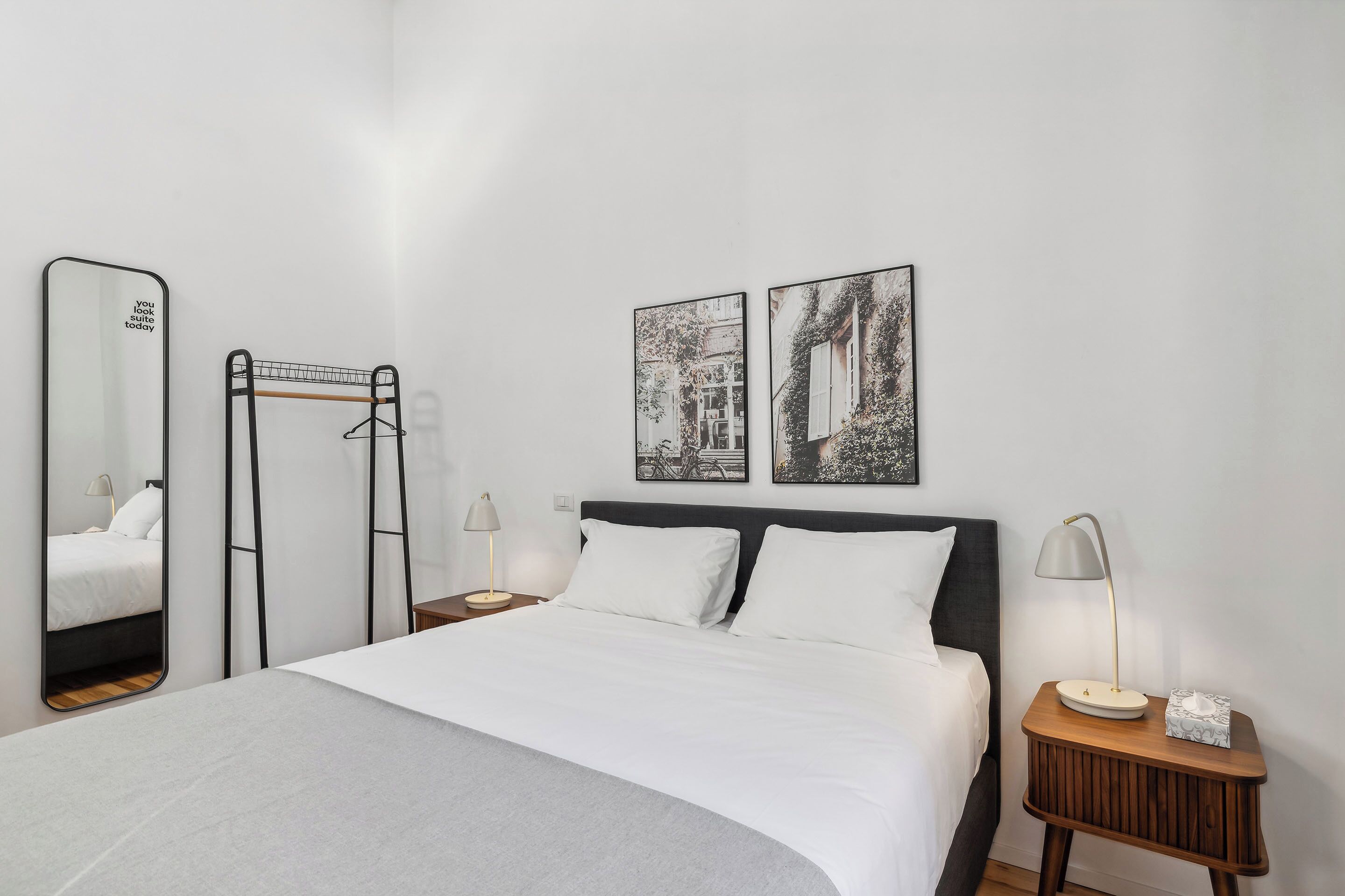 A neatly arranged bedroom with a bed, a bedside table with a lamp, a mirror, and two framed pictures on the wall.