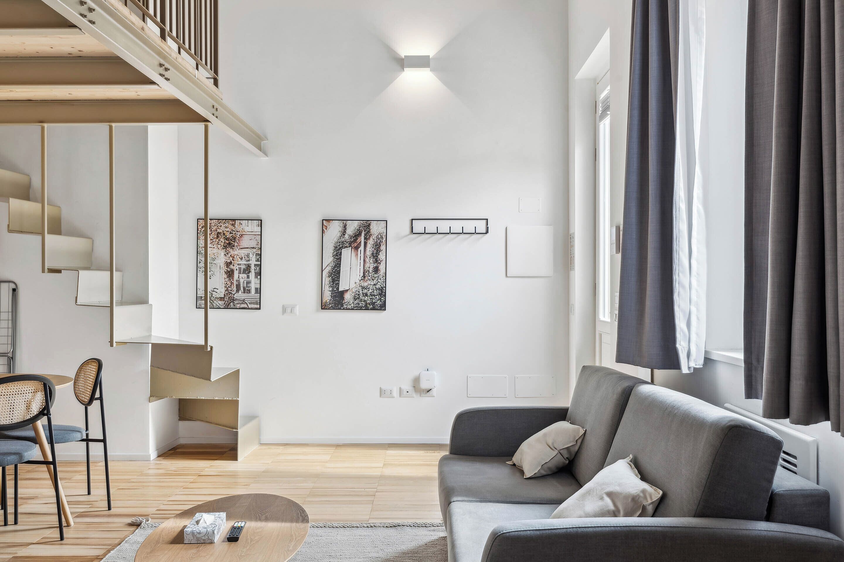 Ein modernes Wohnzimmer mit einem grauen Sofa, einem Holztisch, einer Treppe mit Holzgeländer und zwei gerahmten Kunstwerken an der Wand.