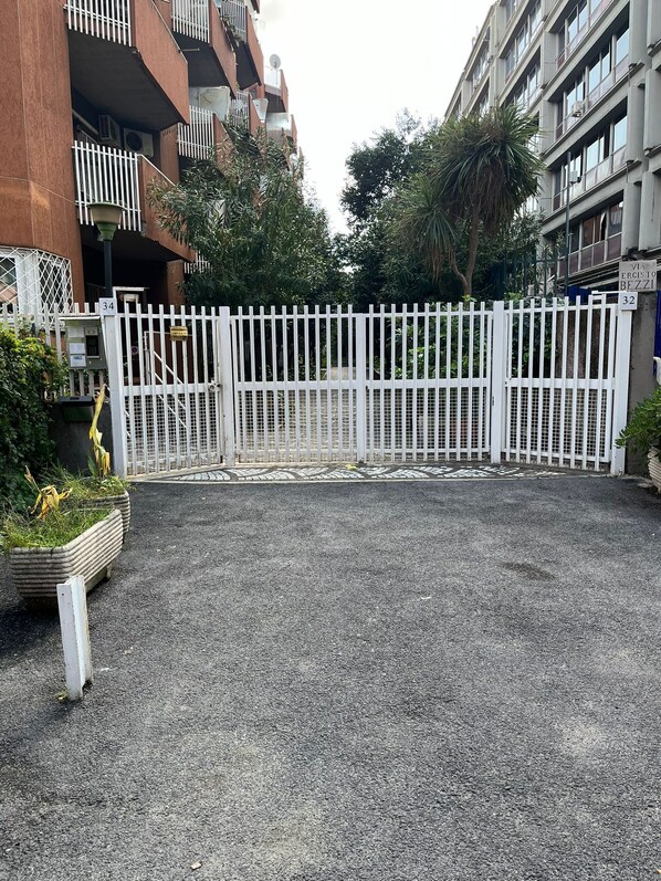 Entrada fechada por portão para um prédio residencial, com acesso por caminho de cascalho e plantas em vasos.