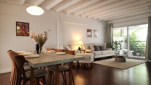 A spacious living area with a dining table set for four, a sofa, and a large window offering a view of greenery outside.
