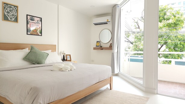 A modern bedroom with a large bed, a bedside table with a lamp, a small shelf with decorative items, and a view of the outdoors through a large window.