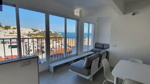 A modern apartment with a balcony overlooking a beach, a sofa, a dining table, and a TV.