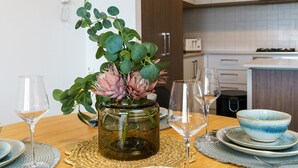 Una mesa de comedor con un florero, dos copas de vino y un tazón azul sobre un mantel tejido.