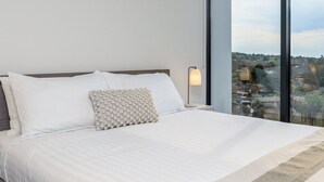 Una cama bien hecha con sábanas blancas y un cojín texturizado, una lámpara de noche y un ventanal con vistas a una zona residencial.