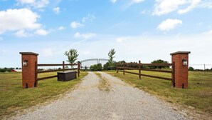 Un chemin de gravier mène à une entrée fermée par un portail, flanquée de piliers en briques et d’une clôture en bois.