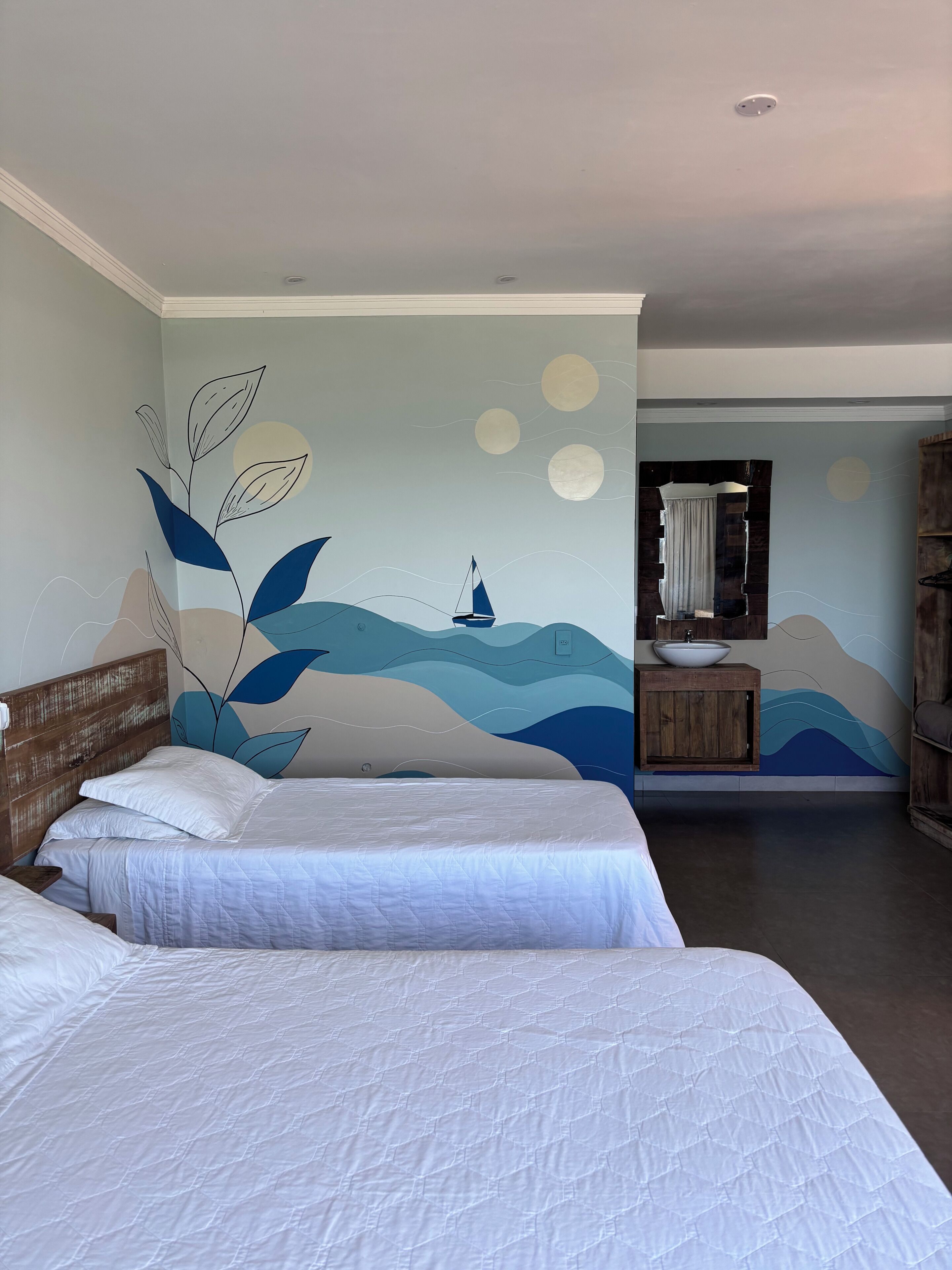 A bedroom with two beds, a wall mural with a sailboat, and a bathroom with a wooden vanity.